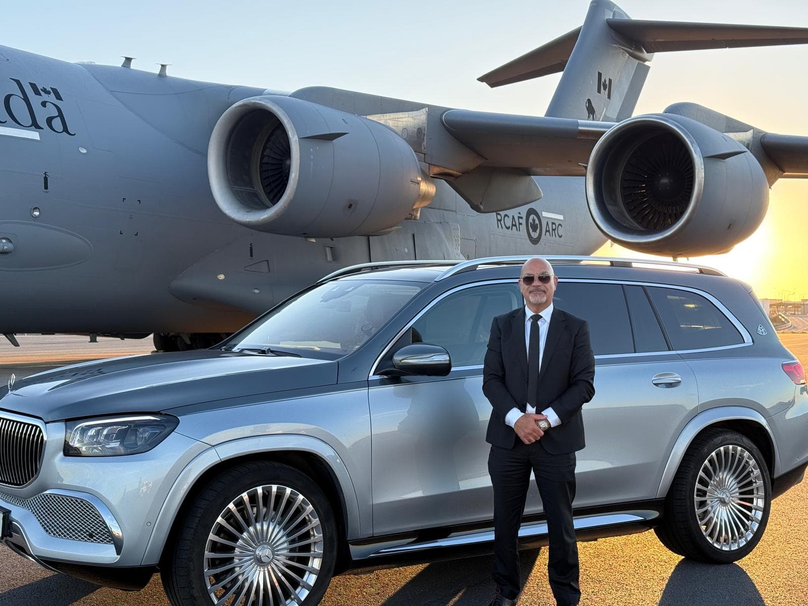Leading Car Service chauffeur in a black suit beside a Mercedes-Maybach GLS and a private jet at sunset